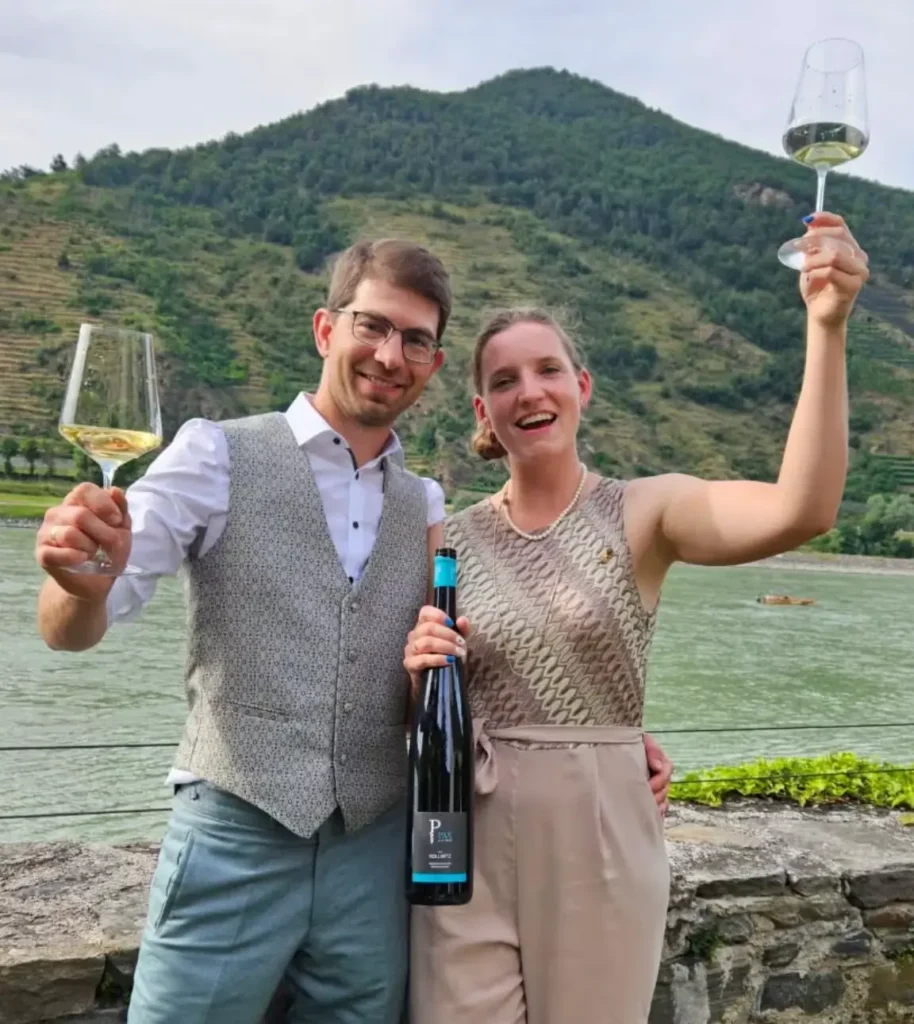 Winzerpaar des Weinguts PAX mit einer Flasche Weißwein vor der Donau in der Wachau.